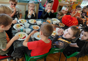 Przedszkolaki przebrane na Halloween siedzą przy stole z halloweenowymi dekoracjami i jedzą drugie śniadanie.