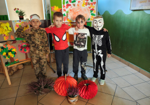 Chłopiec przebrany za rannego żołnierza, chłopiec przebrany za Spidermana, chłopiec przebrany za Jokera i chłopiec przebrany za kościotrupa stoją na podłodze. Przed i za chłopcami jesienna dekoracja.