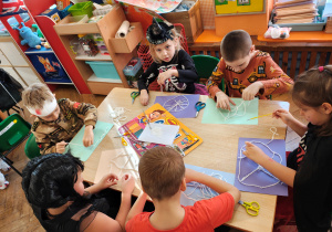 Przedszkolaki przebrane na Halloween siedzą przy stole i wykonują pracę plastyczną - pajęczyny z wełny i pająki z plasteliny.