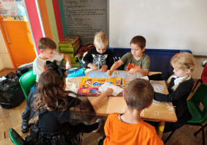 Przedszkolaki przebrane na Halloween siedzą przy stole i wykonują pracę plastyczną - pajęczyny z wełny i pająki z plasteliny.