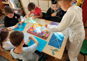 Przedszkolaki przebrane na Halloween siedzą przy stole i wykonują pracę plastyczną - pajęczyny z wełny i pająki z plasteliny.