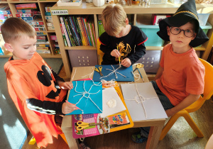 Przedszkolaki przebrane na Halloween siedzą przy stole i wykonują pracę plastyczną - pajęczyny z wełny i pająki z plasteliny.