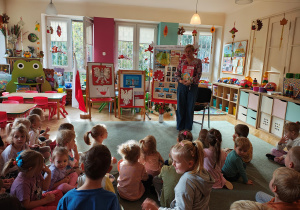 Pani bibliotekarka stoi na dywanie z książką pt. "Legendy Polskie", za nią stoją dekoracje z okazji Święta Niepodległości oraz prace na konkurs "Symbole narodowe", przedszkolaki siedzą na dywanie.