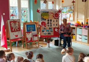 Pani bibliotekarka siedzi na krześle z książką pt. "Legendy Polskie", za nią stoją dekoracje z okazji Święta Niepodległości oraz prace na konkurs "Symbole narodowe", przedszkolaki siedzą na dywanie.