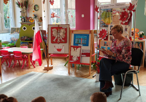 Pani bibliotekarka siedzi na krześle z książką pt. "Legendy Polskie", za nią stoją dekoracje z okazji Święta Niepodległości oraz prace na konkurs "Symbole narodowe", przedszkolaki siedzą na dywanie.