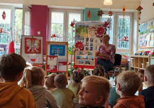 Pani bibliotekarka siedzi na krześle z książką pt. "Legendy Polskie", za nią stoją dekoracje z okazji Święta Niepodległości oraz prace na konkurs "Symbole narodowe", przedszkolaki siedzą na dywanie.