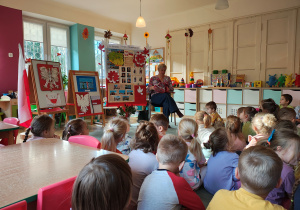 Pani bibliotekarka siedzi na krześle z książką pt. "Legendy Polskie", za nią stoją dekoracje z okazji Święta Niepodległości oraz prace na konkurs "Symbole narodowe", przedszkolaki siedzą na dywanie.