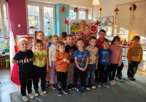Przedszkolaki z grupy II wraz z Panią bibliotekarką stoją na dywanie, część dzieci siedzi, za nimi stoją dekoracje z okazji Święta Niepodległości oraz prace na konkurs "Symbole narodowe".
