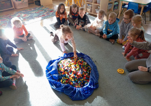 Przedszkolaki siedzą na dywanie. Na środku leży duża torba z klockami LEGO. Dziewczynka bierze klocek z torby.