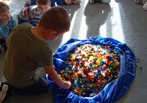 Przedszkolaki siedzą na dywanie. Na środku leży duża torba z klockami LEGO. Chłopiec bierze klocek z torby.