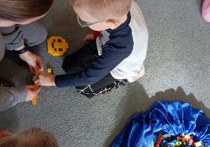 Chłopiec wraz z panią od warsztatów przyczepiają klocka LEGO do budwli. Na dywanie leży duża torba z klockami LEGO.