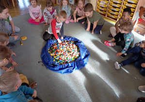 Przedszkolaki wraz z panią od warsztatów siedzą na dywanie. Jeden z chłopców bierze z torby klocka LEGO. Na środku leży duża torba z klockami LEGO.