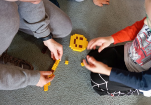 Pani od warsztatów przyczepia klocka LEGO. Przed nią siedzi chłopiec.