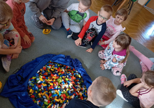 Przedszkolaki wraz z panią od warsztatów siedzą na dywanie. Jeden z chłopców bierze z torby klocka LEGO. Na środku leży duża torba z klockami LEGO.