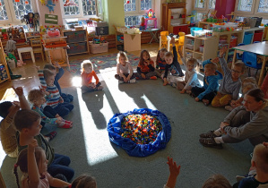 Przedszkolaki wraz z panią od warsztatów siedzą w kole na dywanie. Na środku leży duża torba z klockami LEGO.