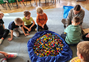 Przedszkolaki wraz z panią od warsztatów siedzą na dywanie. Jeden z chłopców siedzi tyłem. Na środku leży duża torba z klockami LEGO.