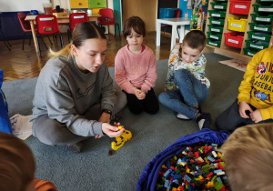 Przedszkolaki wraz z panią od warsztatów siedzą na dywanie. Pani od warsztatów pokazuje klocki. Na dywanie leży duża torba z klockami LEGO.