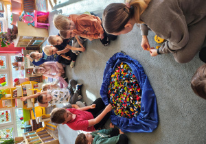 Przedszkolaki wraz z panią od warsztatów siedzą na dywanie. Jeden z chłopców bierze z torby klocka LEGO. Na środku leży duża torba z klockami LEGO.