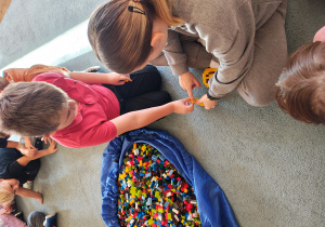 Chłopiec wraz z panią od warsztatów przyczepiają klocka LEGO do budwli. Na dywanie leży duża torba z klockami LEGO.