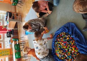 Przedszkolaki wraz z panią od warsztatów siedzą na dywanie. Jeden z chłopców wraz z panią od warsztatów przyczepiają klocka LEGO do budowli. Na środku leży duża torba z klockami LEGO.