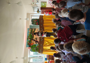 Aktorka w kapeluszu trzymająca kukłę bobra stoi przed scenografią. Przed nią na dywanie siedzą dzieci.