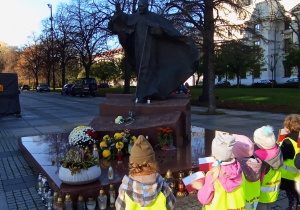 Przedszkolaki w odblaskowych kamizelkach stoją tyłem z flagami Polski przed pomnikiem Jana Pawła II.