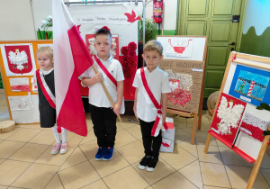 Dwóch chłopców i jedna dziewczynka stoją ubrani na galowo z biało-czerwoną szarfą. Chłopiec stojący w środku trzyma flagę Polski. W tle dekoracja z okazji Święta Niepodległości.