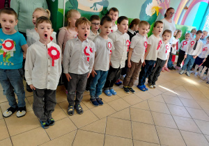 Przedszkolaki wraz z panią, ubrane na galowo z biało-czerwonymi kotylionami stoją na baczność i śpiewają hymn Polski.