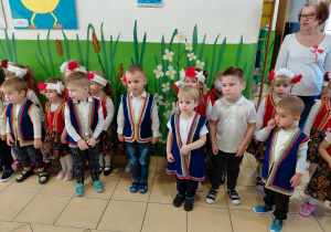 Przedszkolaki wraz z panią, ubrane na galowo w regionalnych kamizelkach, stoją na baczność i śpiewają hymn Polski.