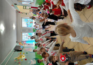 Przedszkolaki wraz z paniami, ubrane na galowo i w regionalnych kamizelkach idą w parach i trójkach po kole, wyciągając ramiona na boki.