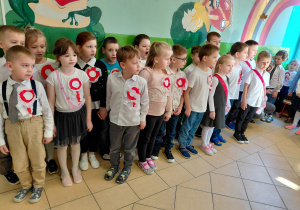 Przedszkolaki stoją ubrane na galowo i w przypiętych biało-czerwonych kotylionach.