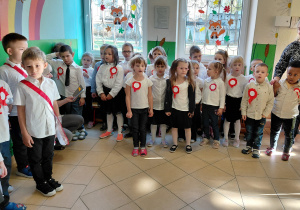 Przedszkolaki stoją ubrane na galowo i w przypiętych biało-czerwonych kotylionach.