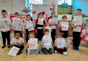 Przedszkolaki ubrane na galowo i z przypiętymi kotylionami stoją trzymając dyplomy konkursu pt. "Symbole narodowe" na tle dekoracji z okazji Święta Niepodległości.