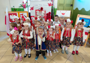 Przedszkolaki z grupy I, ubrane na galowo i w regionalnych kamizelkach stoją na tle dekoracji z okazji Święta Niepodległości.