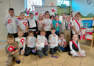 Przedszkolaki z grupy II, ubrane na galowo i z przypiętymi kotylionami stoją na tle dekoracji z okazji Święta Niepodległości.