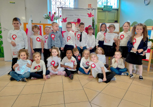 Przedszkolaki z grupy III, ubrane na galowo i z przypiętymi kotylionami stoją na tle dekoracji z okazji Święta Niepodległości.