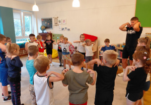 Przedszkolaki w strojach sportowych wraz z trenerem stoją w kole z rękoma na ramionach.