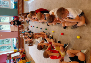 Przedszkolaki siedzą na przeciwko siebie w parach z rękoma na głowach, między nimi leżą piłki.