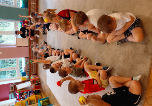 Przedszkolaki siedzą na przeciwko siebie w parach. Niektóre dzieci trzymają piłkę w górze.