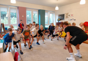 Przedszkolaki w strojach sportowych startują z jednej części dywanu w stronę trenera, ktory stoi w półprzysiadzie na środku dywanu.