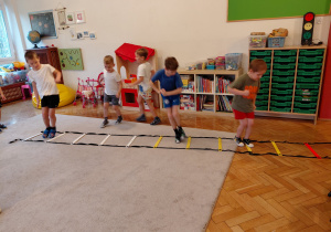 Trzech chłopców skacze między szczebelkami leżącej drabinki, dwóch chłopców biegnie za drabinką.