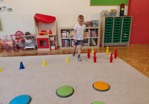 Chłopiec biegnie dookoła ustawionych w kole kręgli, na dywanie leżą sensoryczne koła.