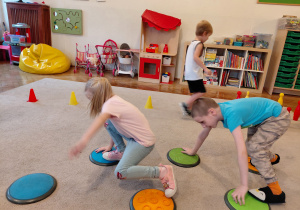 Dziewczynka i chłopiec przechodzą po sensorycznych kołach. Z tyłu między pachołkami biegnie chłopiec.