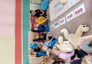 Przedszkolaki siedzą z pluszakami przed kartonikami z numerami alarmowymi. Niektóre dzieci podnoszą rękę. Na dywaniku leży manekin do ćwiczeń resuscytacji.