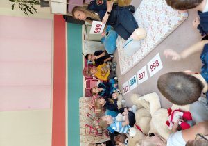 Przedszkolaki siedzą z pluszakami przed kartonikami z numerami alarmowymi. Na dywaniku siedzi strażniczka miejska trzymająca tabliczkę z numerem alarmowym pogotowia ratunkowego. Przed nią leży manekin do ćwiczeń resuscytacji.