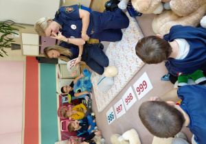 Przedszkolaki siedzą z pluszakami przed kartonikami z numerami alarmowymi. Na dywaniku siedzą strażniczki miejskie, jedna z nich trzyma tabliczkę z numerem alarmowym 112. Przed nimi leży manekin do ćwiczeń resuscytacji.