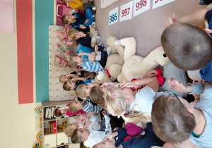 Przedszkolaki siedzą z pluszakami przed kartonikami z numerami alarmowymi. Niektóre dzieci wskazują palcami na swoje oczy.