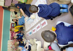 Przedszkolaki siedzą z pluszakami przed kartonikami z numerami alarmowymi. Na dywaniku siedzi strażniczka miejska trzymająca nożyczki. Przed nią leży manekin do ćwiczeń resuscytacji.