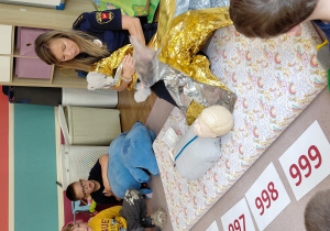 Na dywanie siedzi strażniczka miejska owijająca misia w koc termiczny. Przed nią leży manekin do ćwiczeń resuscytacji. Za dywanem siedzi dwóch chłopców.