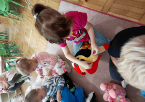 Przedszkolaki siedząc przyklejają plastry pluszakom.